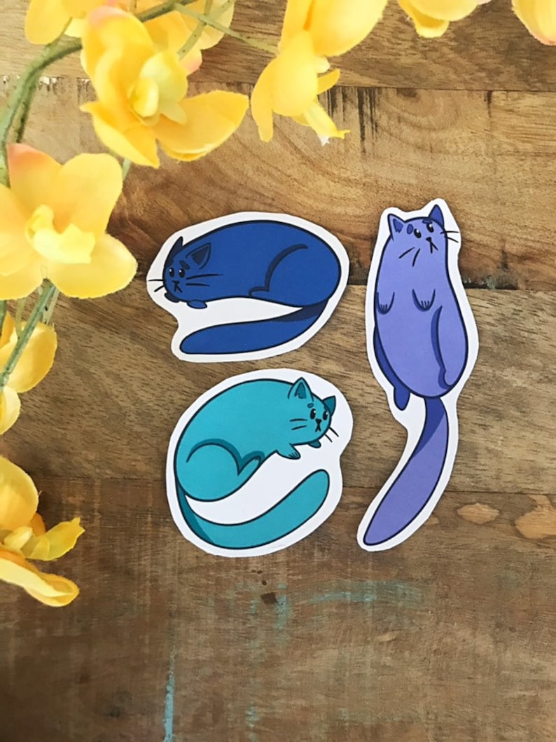 K&ouml;nnte beinhalten: Drei Katzenaufkleber mit verschiedenen Posen. Die Katze links ist blau und zusammengerollt. Die Katze in der Mitte ist t&uuml;rkis und zusammengerollt. Die Katze rechts ist lila und steht auf.