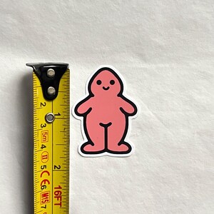 May include: A pink sticker with a cartoon figure design. The figure has a rounded head, a smiling face, and a body with rounded arms and legs. The sticker is approximately 2 inches tall.