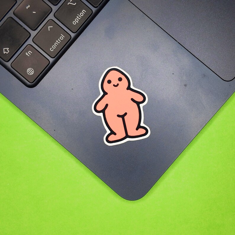 May include: A peach-colored sticker with a cartoon figure design. The sticker has a black outline and a smiling face. It is placed on a dark gray laptop, with a bright green background.