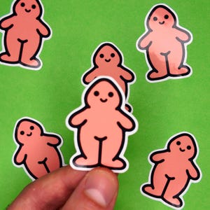 May include: A collection of peach-colored stickers with a cartoonish figure design. Each sticker features a smiling face and a simple body outline with black detailing. The stickers are on a green background.