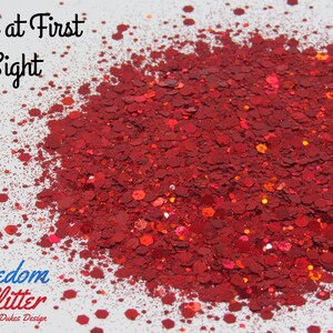 May include: A pile of red glitter with various shapes and sizes. The glitter is scattered on a white surface. The text "Love at First Sight" is at the top, and "Freedom Glitter by Dukes Design" is at the bottom.