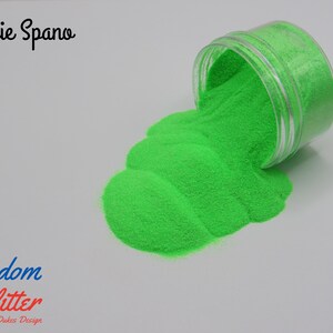 May include: A clear plastic jar tipped over, spilling bright green glitter onto a white surface. The glitter forms a textured, flowing pattern. Text on the image reads "Jessie Spano" and "Freedom Glitter By Dukes Design".