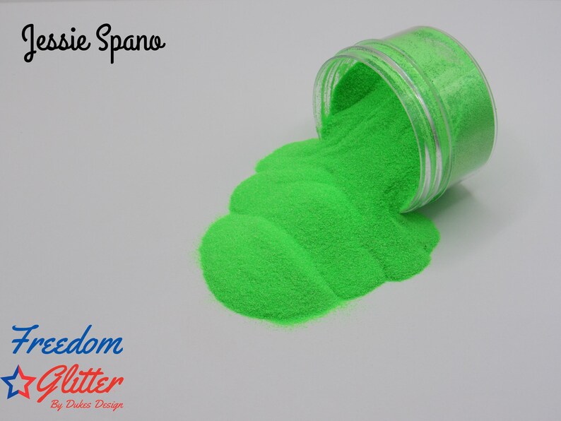 May include: A clear plastic jar tipped over, spilling bright green glitter onto a white surface. The glitter forms a textured, flowing pattern. Text on the image reads "Jessie Spano" and "Freedom Glitter By Dukes Design".