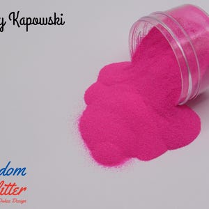 May include: A clear plastic jar tipped over, spilling bright pink glitter onto a white surface. The text "Kelly Kapowski" is above the glitter, and "Freedom Glitter" is below. The glitter is a vibrant, eye-catching color.