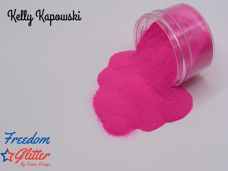 May include: A clear plastic jar tipped over, spilling bright pink glitter onto a white surface. The text "Kelly Kapowski" is above the glitter, and "Freedom Glitter" is below. The glitter is a vibrant, eye-catching color.