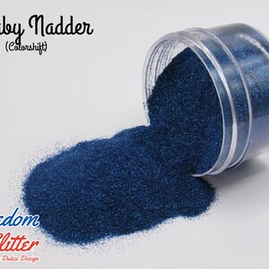 May include: A clear plastic jar tipped over, spilling out a pile of dark blue glitter. The glitter is labeled "Baby Nadder (Colorshift)" with the "Freedom Glitter" logo below. The glitter has a shimmering, metallic appearance.