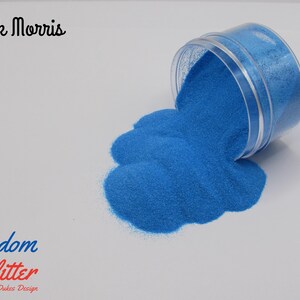 May include: A clear plastic container tipped over, spilling bright blue glitter onto a white surface. The text "Freedom Glitter" and "By Dukes Design" is visible. The name "Zack Morris" is also written in cursive.