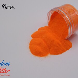 May include: A clear plastic jar tipped over, spilling bright orange glitter onto a white surface. The glitter forms a small pile. Text in the image reads "G.C. Slater" and "Freedom Glitter By Dukes Design".