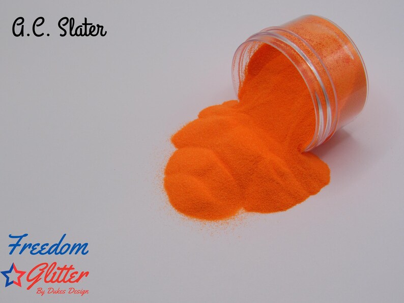 May include: A clear plastic jar tipped over, spilling bright orange glitter onto a white surface. The glitter forms a small pile. Text in the image reads "G.C. Slater" and "Freedom Glitter By Dukes Design".