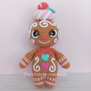 Puede incluir: Un juguete de jengibre de crochet con un sombrero de remolino rosa y blanco, ojos marrones, una nariz roja y una corbata de lazo roja. El juguete tiene detalles de glaseado blanco y corazones rosas, azules y blancos en su cuerpo. El juguete está de pie sobre una superficie blanca y tiene el texto "@MISSDAISYHANDMADE" en la parte inferior.