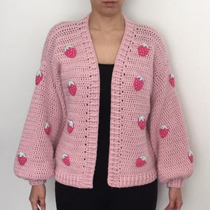 Peut inclure: Un cardigan en crochet rose avec des manches longues et un col en V. Le cardigan est décoré de fraises brodées.