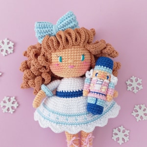 Pode incluir: Uma boneca de crochê com cabelo castanho, um laço azul e um vestido branco segura uma boneca quebra-nozes de crochê. As bonecas estão em um fundo rosa com flocos de neve brancos.