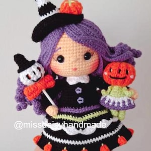 May include: A handmade crochet doll in a witch costume, featuring a black dress with white trim, purple hair, and a witch hat. The doll holds a pumpkin-headed doll and a small doll with a witch hat. The doll has the text "@missdaisyhandmade".