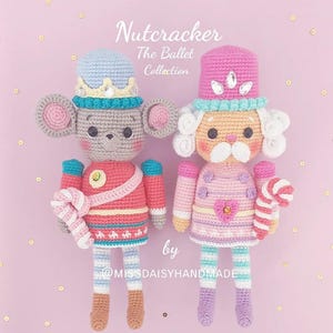 May include: Two crocheted nutcracker dolls, one a mouse and one a traditional nutcracker. The mouse nutcracker is wearing a pink and red uniform with a candy cane. The traditional nutcracker is wearing a pink and purple uniform with a candy cane. Both dolls have a white and blue striped pattern on their legs. The dolls are on a pink background with gold glitter.