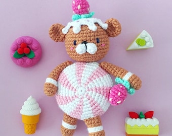 May include: A crocheted teddy bear wearing a pink and white candy-themed outfit with a strawberry on top of its head. The bear is surrounded by small, colourful, crocheted food items.
