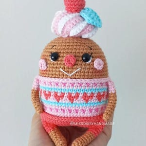 May include: A crocheted amigurumi toy shaped like a gingerbread person wearing a pink and blue sweater with hearts and a pink and white swirl hat with a red cherry on top. The toy has a smiling face with black eyes and a red nose.