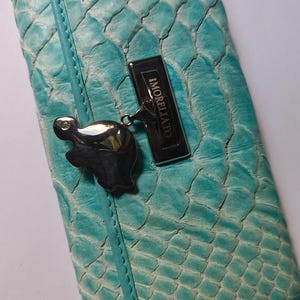 May include: A turquoise leather wallet with a reptile scale pattern. The wallet has a silver-tone clasp with a decorative charm and the brand name "MORELLATO" on a metal plate.