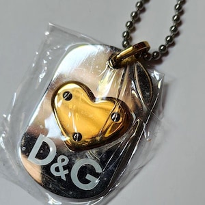 May include: A silver-coloured dog tag pendant with a gold heart-shaped accent. The pendant has the letters "D&G" in white. The pendant is attached to a silver ball chain necklace.