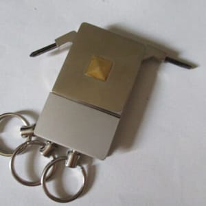 Puede incluir: Un llavero plateado con una pequeña herramienta multiusos adjunta. La herramienta multiusos tiene un pequeño cuadrado dorado en la parte superior y una cuchilla plateada que se pliega.