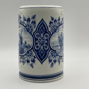 May include: A white ceramic mug with a blue and white Delft design. The mug features a windmill, a horse-drawn carriage, and floral patterns.