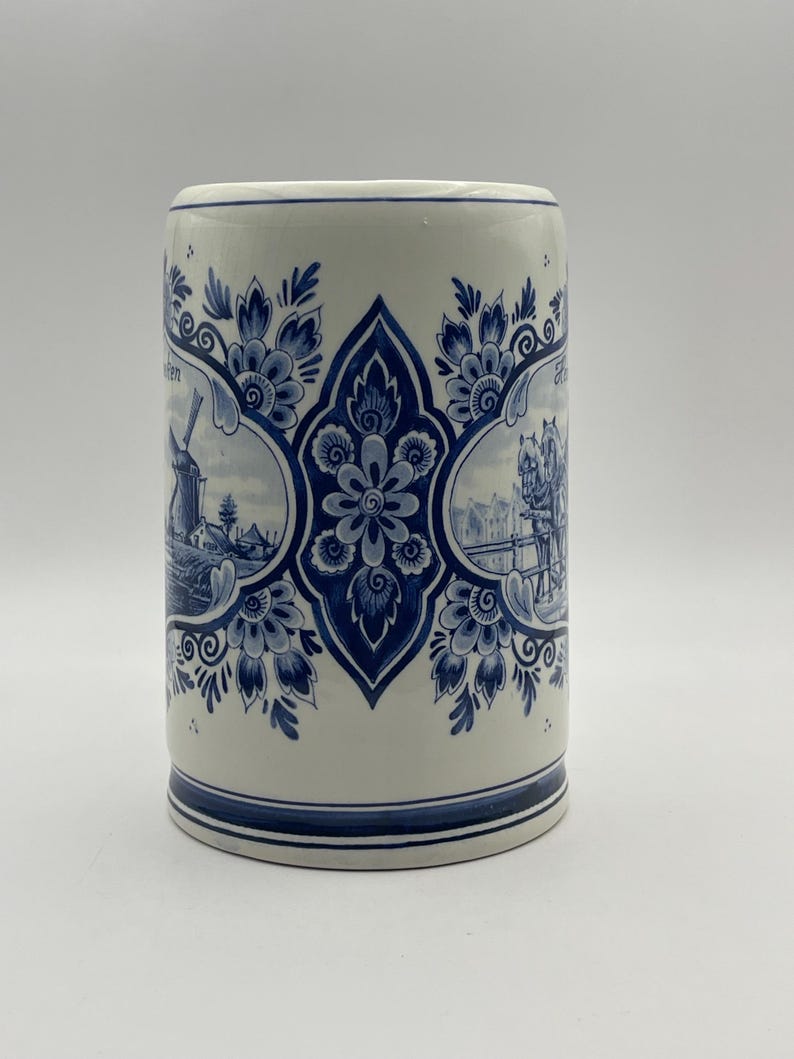 May include: A white ceramic mug with a blue and white Delft design. The mug features a windmill, a horse-drawn carriage, and floral patterns.