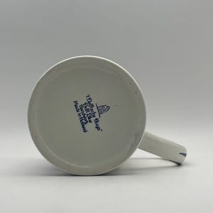 May include: White ceramic mug with a blue Delft Blue design. The mug has a blue handle and a blue inscription that reads "Delftche Huys. Delft Blue Handwork Made in Holland."