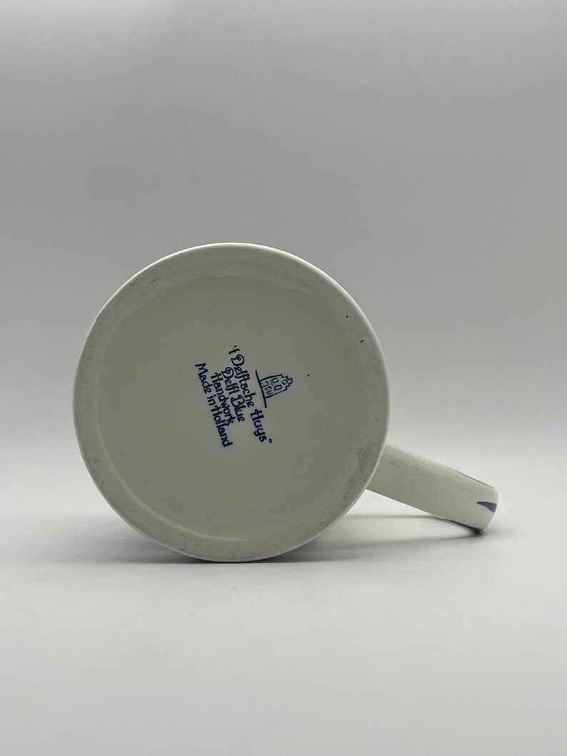 May include: White ceramic mug with a blue Delft Blue design. The mug has a blue handle and a blue inscription that reads "Delftche Huys. Delft Blue Handwork Made in Holland."