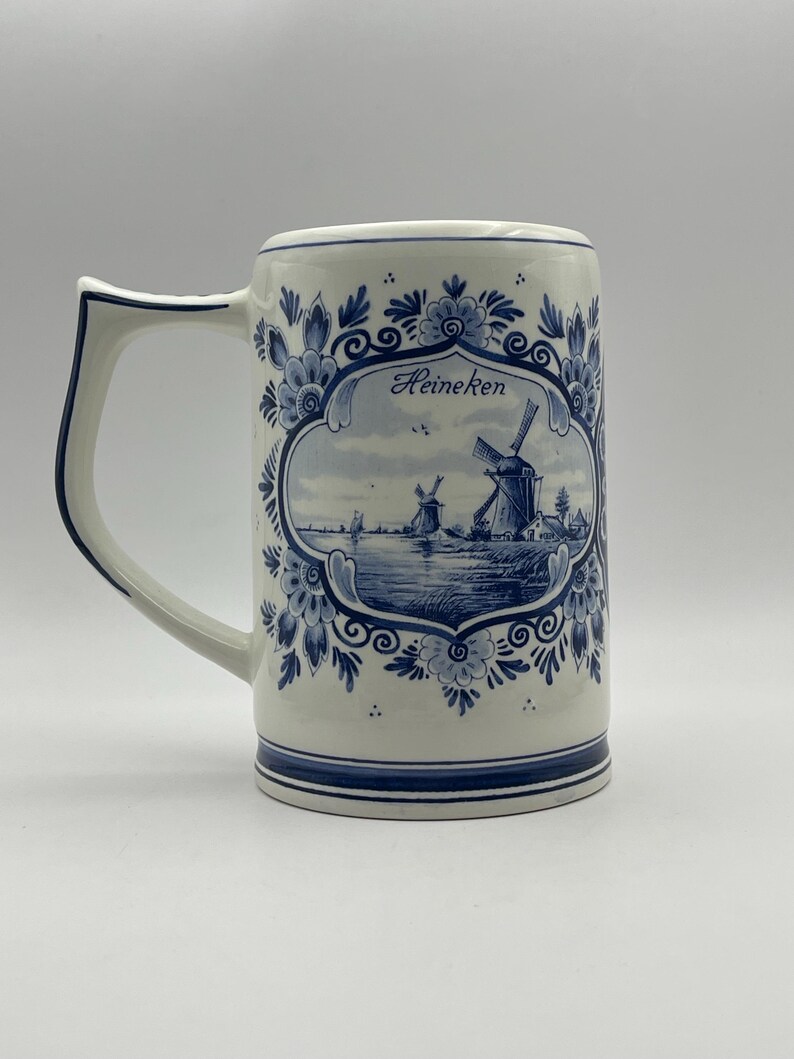 May include: A white ceramic beer stein with a blue and white Delft design featuring windmills and the word "Heineken".