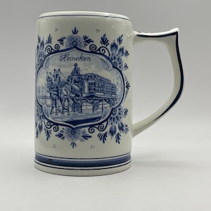 May include: A white ceramic beer stein with a blue and white Delft design. The stein features a horse-drawn carriage with the word "Heineken" on it.