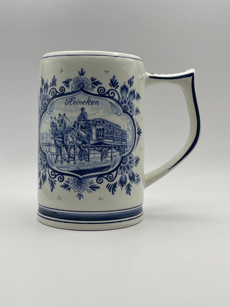 May include: A white ceramic beer stein with a blue and white Delft design. The stein features a horse-drawn carriage with the word "Heineken" on it.