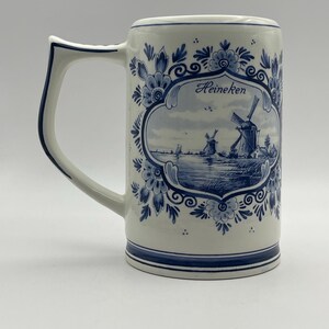 May include: A white ceramic beer stein with a blue and white Delft design. The stein features a windmill scene with the word "Heineken" in a blue oval.