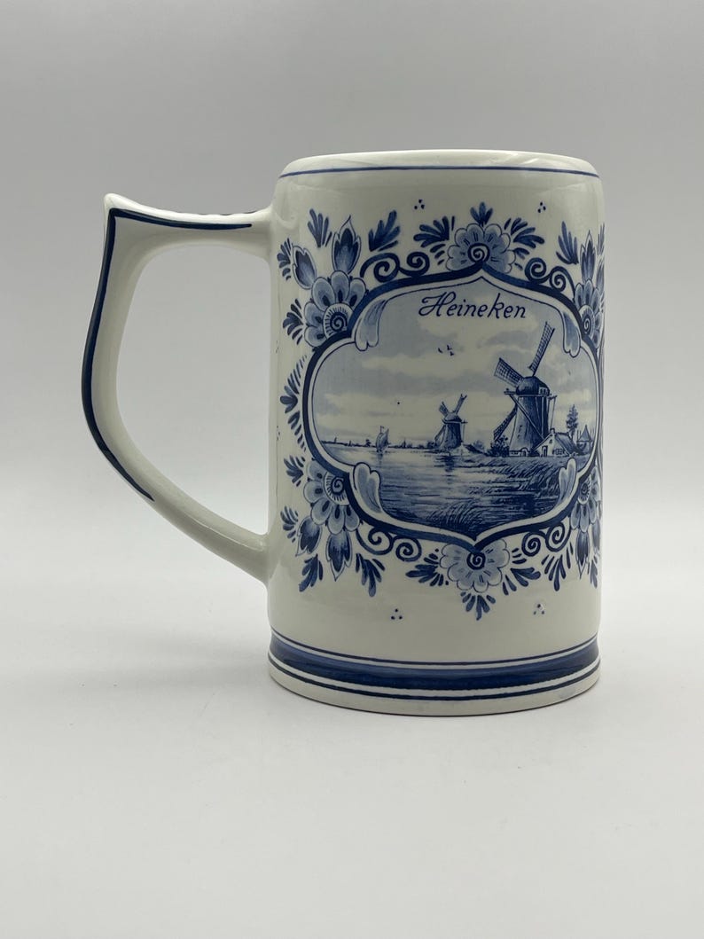 May include: A white ceramic beer stein with a blue and white Delft design. The stein features a windmill scene with the word "Heineken" in a blue oval.