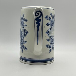 May include: A white ceramic mug with a blue and white Delft design. The mug has a handle with a blue swirl design. The mug features a scene of a Dutch canal with houses and boats.