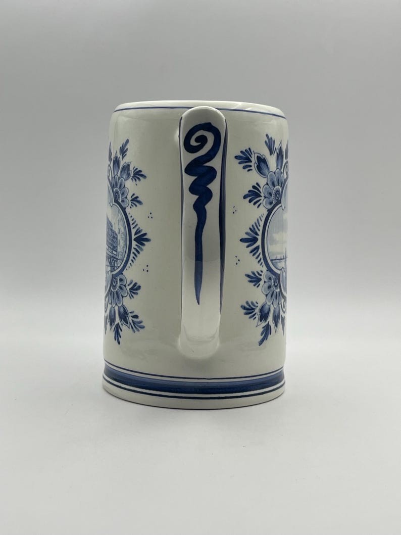 May include: A white ceramic mug with a blue and white Delft design. The mug has a handle with a blue swirl design. The mug features a scene of a Dutch canal with houses and boats.