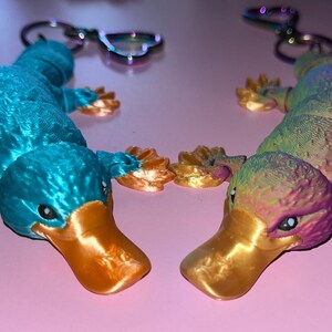 May include: Two platypus keychains, one is teal blue with gold accents and the other is a rainbow of colors with gold accents.