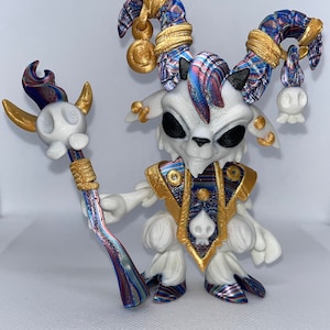 May include: A white, 3D printed figurine of a goat-like creature with gold and blue horns, wearing a gold and blue patterned robe and holding a staff with a skull at the top.