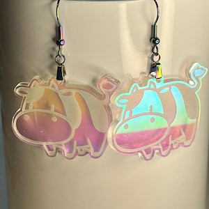 Puede incluir: Un par de pendientes de vaca iridiscentes. Los pendientes están hechos de acrílico transparente y presentan un diseño de vaca de dibujos animados. Las vacas son rosas y azules con un acabado iridiscente arcoíris.