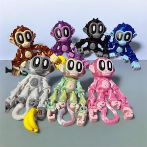 May include: A collection of colorful articulated monkey figurines in various colors, including brown, purple, black, blue, white, green, and pink. Each monkey has a unique design with large, oval eyes and a flexible body. A yellow banana is included.