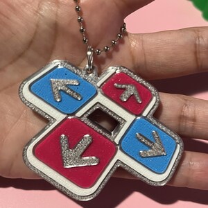 May include: A silver-colored keychain pendant featuring a dance pad design. The pendant has four colored squares: red and blue, each with a silver arrow. The pendant is attached to a silver ball chain.