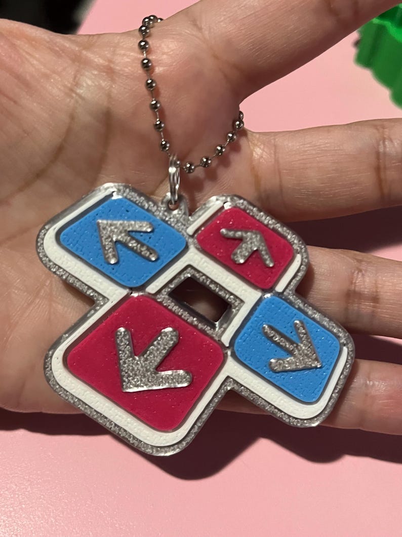May include: A silver-colored keychain pendant featuring a dance pad design. The pendant has four colored squares: red and blue, each with a silver arrow. The pendant is attached to a silver ball chain.