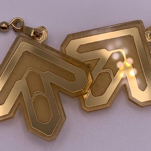 Puede incluir: Un par de pendientes de color dorado con un diseño geométrico. Los pendientes están hechos de un material transparente con un acabado dorado. Los pendientes tienen forma de flechas que apuntan hacia arriba.