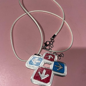 May include: A necklace featuring a pendant shaped like a dance pad. The pendant has four directional buttons in red and blue, each with a silver arrow. The necklace has a white cord and a silver clasp.