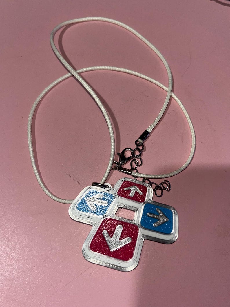 May include: A necklace featuring a pendant shaped like a dance pad. The pendant has four directional buttons in red and blue, each with a silver arrow. The necklace has a white cord and a silver clasp.