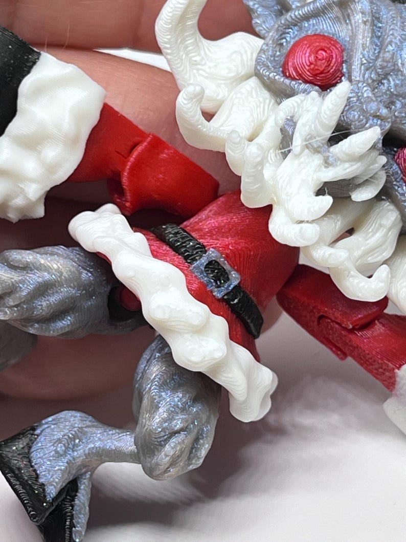 May include: A close-up of a 3D-printed Santa Claus figurine. The figure is predominantly red, white, and gray, with intricate details. The Santa's coat has white trim, and a black belt. The figure's legs and arms are gray.