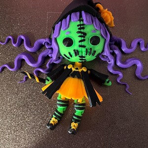 May include: A green and purple witch doll with stitched seams, a black hat, and a yellow and orange dress. The doll has long purple hair and is wearing black and green striped stockings.
