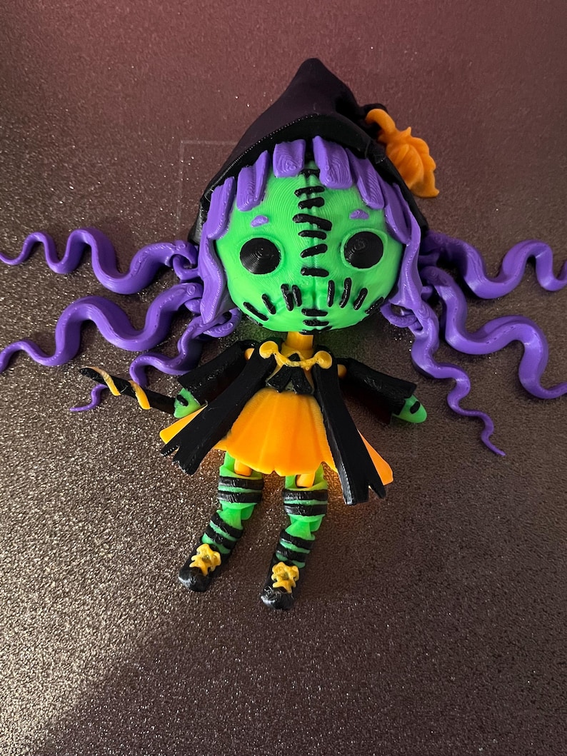 May include: A green and purple witch doll with stitched seams, a black hat, and a yellow and orange dress. The doll has long purple hair and is wearing black and green striped stockings.