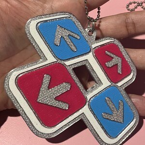 May include: A colorful keychain featuring a dance pad design. The keychain has a silver glitter border and a white outline. The arrows are silver, set against blue and red squares. A silver ball chain is attached.