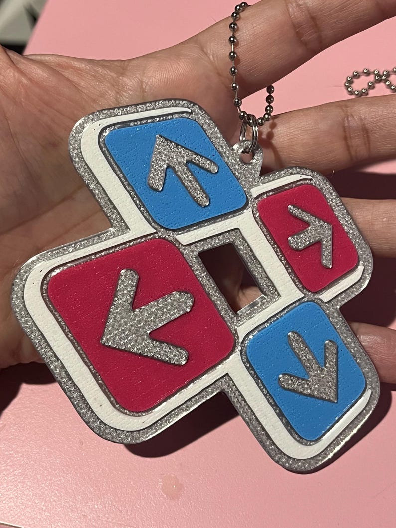 May include: A colorful keychain featuring a dance pad design. The keychain has a silver glitter border and a white outline. The arrows are silver, set against blue and red squares. A silver ball chain is attached.