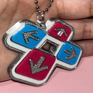 May include: A silver-colored pendant necklace featuring a cross-shaped design with blue and pink squares. Each square has a silver arrow pointing towards the center. The pendant is attached to a silver ball chain.