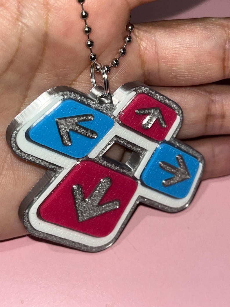 May include: A silver-colored pendant necklace featuring a cross-shaped design with blue and pink squares. Each square has a silver arrow pointing towards the center. The pendant is attached to a silver ball chain.
